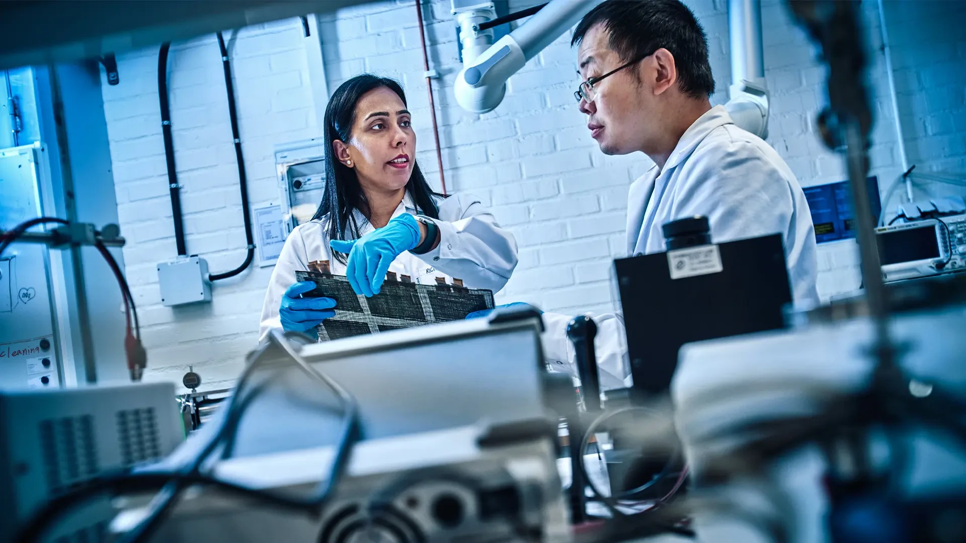 Two researchers in white lab coats examining a carbon fiber composite in a high-tech laboratory focused on structural batteries