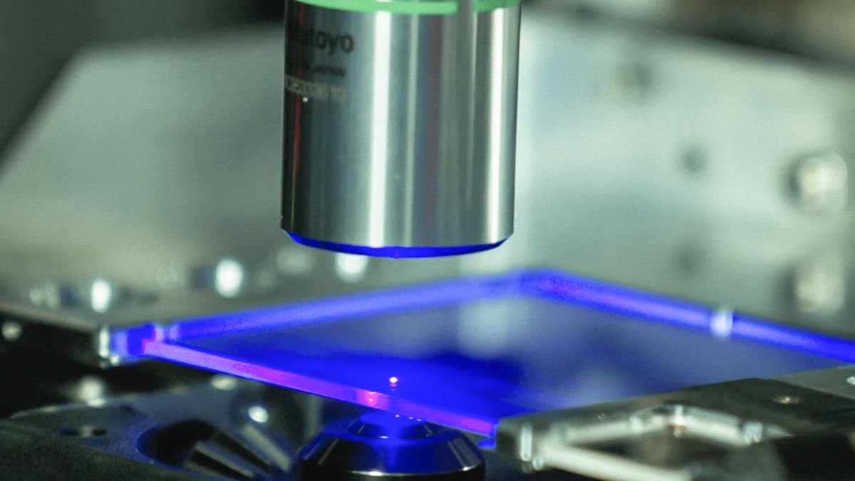 Macro close-up of a femtosecond laser beam writing data on a Microsoft Project Silica quartz glass plate, glowing blue during the process.